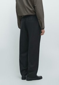 Schwarze, weit geschnittene Hosen mit glatter Textur, elastischem Bund und minimalistischem Design. Von hinten gezeigt, kombiniert mit schwarzen Schuhen.