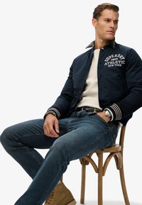 Chaqueta bomber naval con logo, letras blancas y puños a rayas; suéter crema debajo, jeans azules y botas marrones con un reloj.