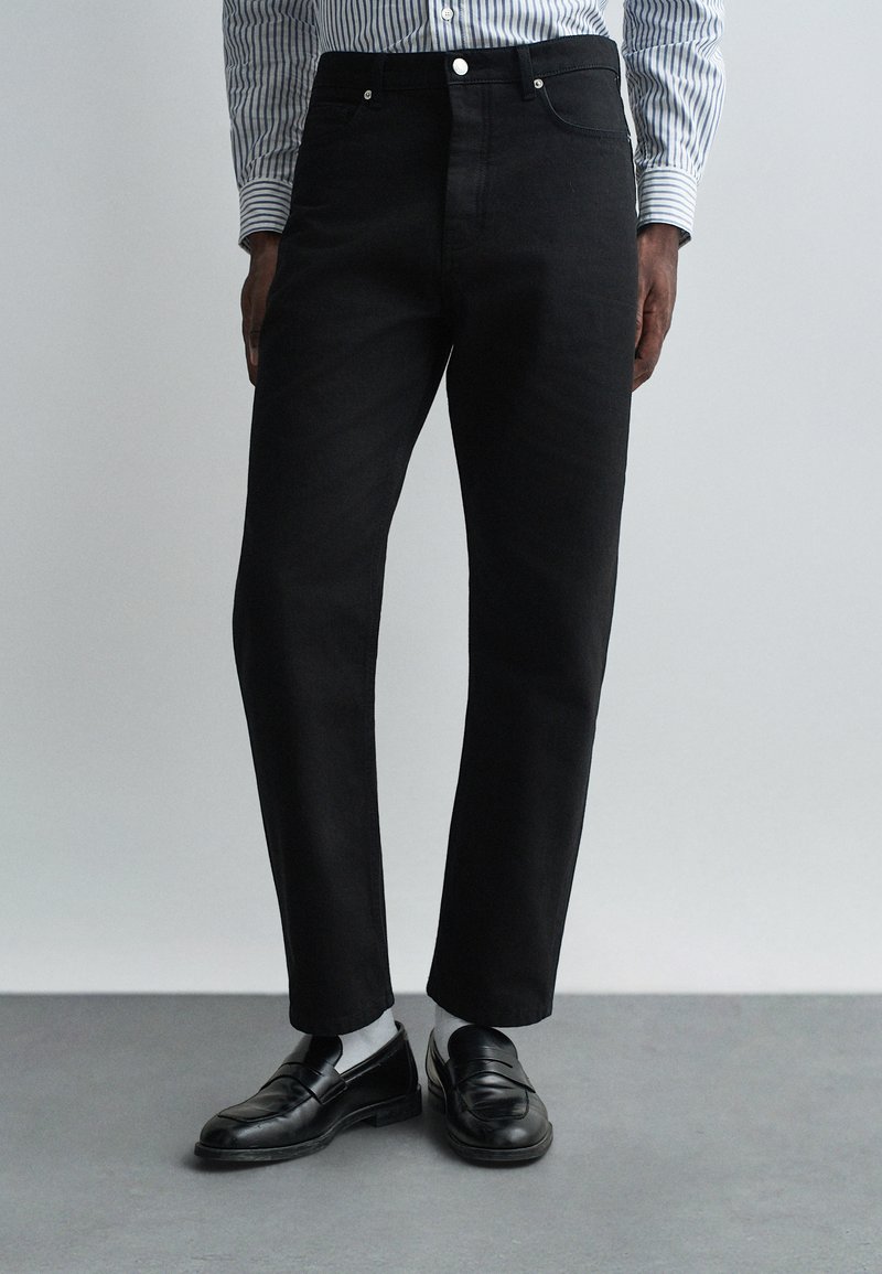 Pantaloni negri din denim cu croială dreaptă, având un design cu cinci buzunare și accesorii din argint. Purtați cu loaferi negri și ciorapi gri.