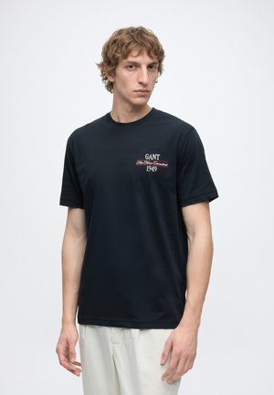Jeune homme aux cheveux châtain clair porte un t-shirt noir GANT avec un texte blanc et un pantalon blanc cassé, debout devant un fond clair uni.