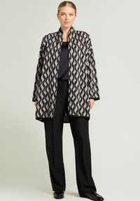 Lange, openvallende cardigan met een golfpatroon, uitgevoerd in crème- en zwarte kleuren met een gestructureerde afwerking. Combinatie met zwarte broek en schoenen.