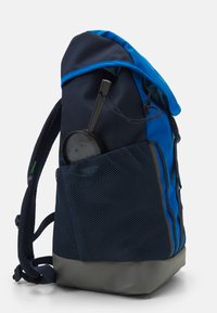 Mochila de tela azul y marino con base gris, bolsillo lateral de malla, correas ajustables y un cierre de cordón en la parte superior.