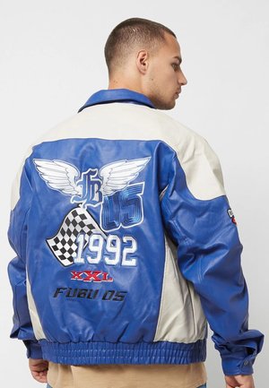 Junger Mann trägt eine blau-weiße Rennjacke mit Flügel, Schachflagge und 1992-Stickerei auf dem Rücken.