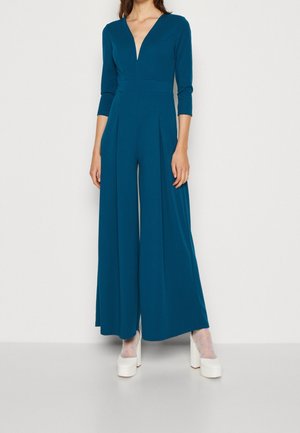 Tyrkysový overal s hlubokým V výstřihem, dlouhými rukávy, širokými nohavicemi a překládaným předním dílem. Doplňený bílými platformovými podpatky. Hladká textilie.