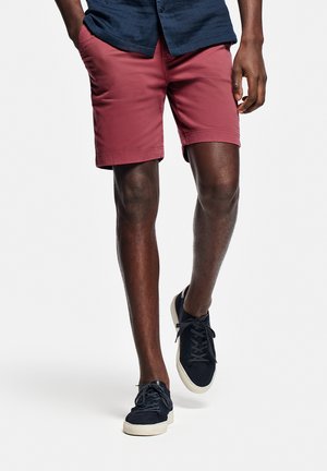 Man draagt donkerrode korte broek, marineblauw shirt en zwarte sneakers, loopt met één hand in de zak tegen een witte achtergrond.