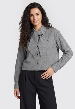 Donna con lunghi capelli ricci che indossa una camicetta a quadri bianco e nero con un grande fiocco e pantaloni neri, sorride leggermente, mano sui fianchi.
