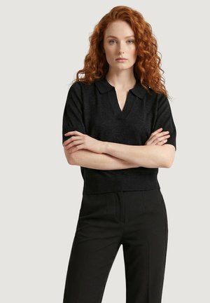 Donna dai capelli rossi e pelle chiara indossa una maglietta nera a maniche corte con colletto e pantaloni neri, in piedi con le braccia incrociate davanti a uno sfondo semplice.