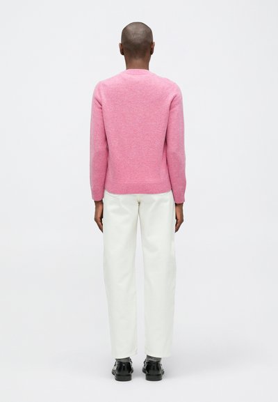 Osoba z ogoloną głową, ubrana w różowy sweter, białe spodnie, szare skarpety i czarne buty, stojąca tyłem na tle białym.