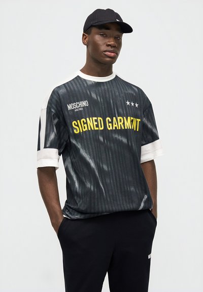 Νέος άνδρας με μαύρο καπέλο, μαύρο πουκάμισο με ρίγες και το κείμενο "SIGNED GARMENT" και μαύρο παντελόνι, με τα χέρια στις τσέπες, μπροστά σε απλό φόντο.