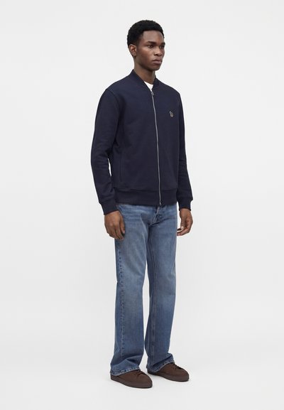 Homme portant une veste zippée bleu marine, un jean bleu clair et des chaussures marron, debout devant un fond blanc uni.