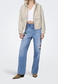 Beige cropped jacket with a hood, paired with blue wide-leg jeans featuring floral embroidery. Model wears pointed tan ankle boots.