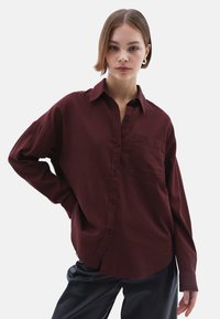 Camicia bordeaux con bottoni realizzata in tessuto morbido, dotata di tasca sul petto, vestibilità ampia e maniche lunghe. Indossata con pantaloni neri.