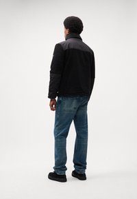 Giacca in pile e nylon nera con colletto alto, abbinata a jeans di denim blu e scarpe con tacco nere, vista da dietro.