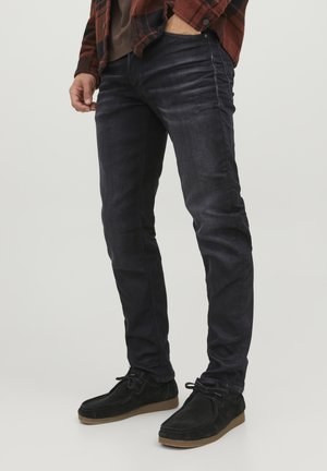 Jeans de mezclilla oscura con un corte ajustado, con detalles sutiles de desgaste y arrugas. Combinados con zapatos de ante negros sobre un fondo claro.