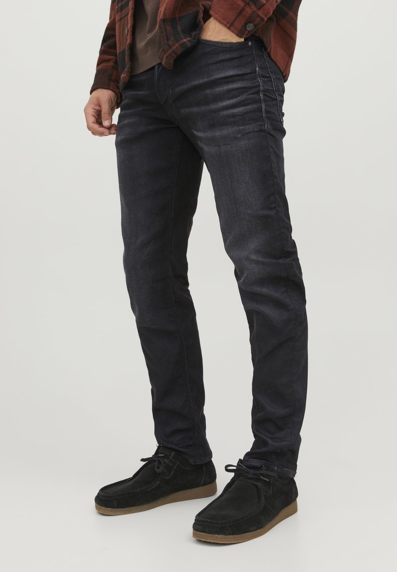 Jeans de mezclilla oscura con un corte ajustado, con detalles sutiles de desgaste y arrugas. Combinados con zapatos de ante negros sobre un fondo claro.
