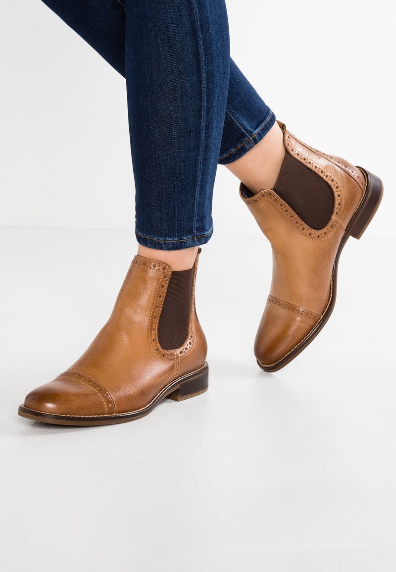 Bottines Chelsea en cuir fauve avec détails brogue portées avec un jean skinny bleu foncé, vues sur un fond de sol blanc.