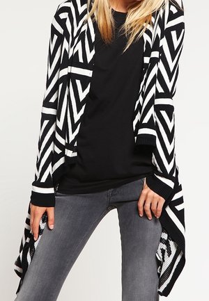Personne portant une chemise noire, un jean gris et un long cardigan à motifs zigzag noir et blanc avec un ourlet asymétrique.