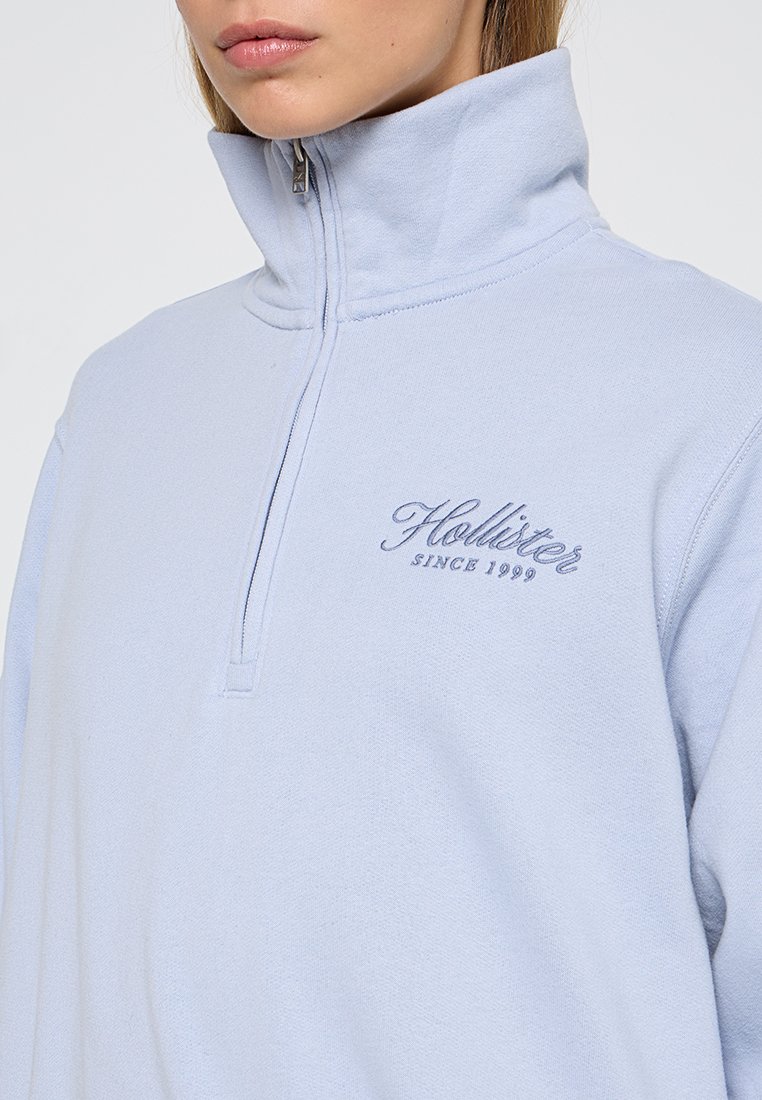 Pull en bleu clair avec col haut, fermeture zippée sur la moitié, et logo brodé. Matériau en coton doux avec une surface légèrement texturée.