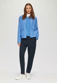 Lichtblauwe ritsjack met twee voorkruiszakken, gedragen over een bijpassende blauwe top, gecombineerd met donker tapse broek en witte sneakers.