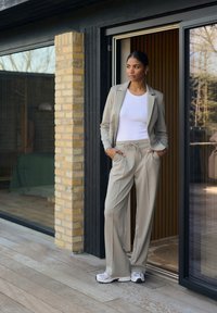 Blazer beige su una maglietta bianca, abbinato a pantaloni coordinati. Scarpe da ginnastica bianche, texture liscia, in piedi su una decking in legno.