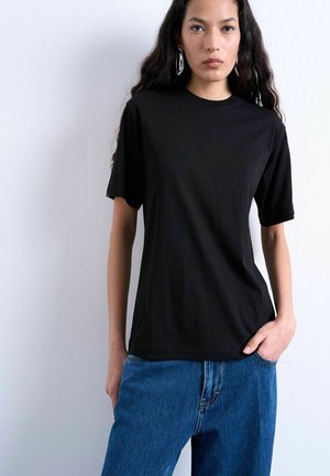 Young woman with long wavy hair wearing large geometric earrings, a black short-sleeve shirt, and blue jeans with one hand in pocket.