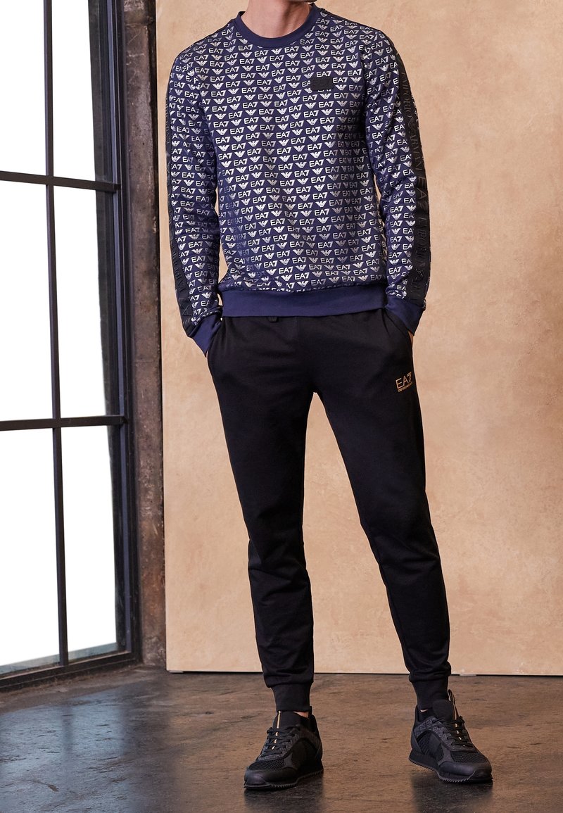 Marineblauer Sweatshirt mit weißem Logomuster, langen Ärmeln und gerippten Bündchen, kombiniert mit schwarzen Jogginghosen und schwarzen Sportschuhen.