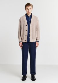 Beige gerippter Cardigan mit fünf Knöpfen, getragen über einem blauen Denim-Hemd, kombiniert mit blauen Hosen und schwarzen Schuhen.