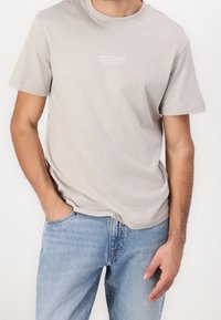 Světle šedé bavlněné tričko s kulatým výstřihem a krátkými rukávy, s jemným bílým logem na hrudi, v kombinaci s modrými džínami.