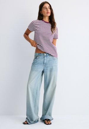 Femme portant un t-shirt rayé violet et blanc, un jean bleu clair à jambes larges et des sandales noires, debout devant un fond blanc.