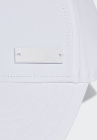 Gorra blanca con una textura de tela suave, visera curvada y una etiqueta blanca con la marca que presenta detalles en relieve, asegurada con dos orificios.
