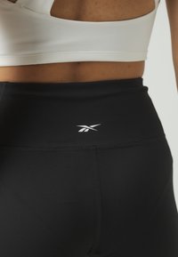 Crne sportske tajice s visokim strukom, s bijelim logotipom na stražnjoj strani. Mekani, rastezljivi materijal s glatkom teksturom.