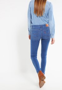 Jeans de mezclilla azul ajustados con una cintura alta, bolsillos traseros y una etiqueta de marca de cuero. Combinados con una camisa azul clara y botas de tacón marrones.