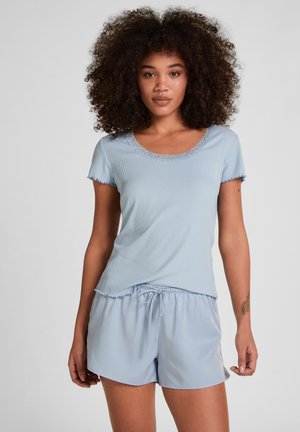 Jonge vrouw met krullend haar, gekleed in een lichtblauw shirtje met korte mouwen en kantafwerking, gecombineerd met bijpassende short met aantrekkoord, tegen een effen achtergrond.