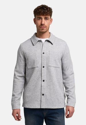 Młody mężczyzna ubrany w jasnoszarą pikowaną kurtkę z czarnymi guzikami, na białej koszuli i niebieskich dżinsach, stojący na jednolitym tle.