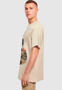 Beige bomulls t-skjorte med korte ermer, med et fargerikt grafisk trykk av palmer og en studiovaske på forsiden.