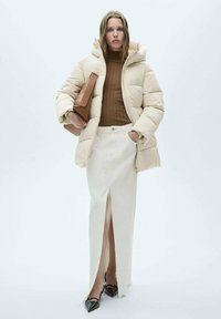 Beige pufferjack met capuchon, bruine geribbelde trui, witte rok met een split aan de voorkant, zwarte puntige hakken en een bruine handtas.