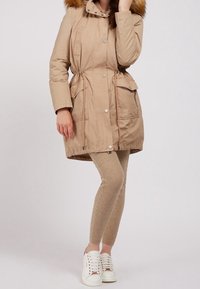 Kvinde iført beige parka med pelskantet hætte, matchende strikkede leggings og hvide sneakers, stående mod en ensfarvet baggrund.