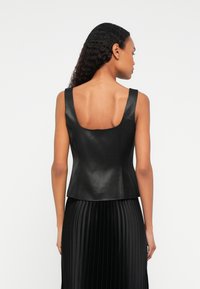 Vrouw met lang, krullend haar, gekleed in een mouwloos zwart rib-top en een zwarte plissé rok, met de rug naar de camera gericht tegen een effen achtergrond.