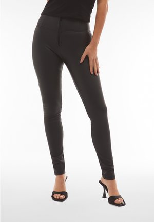 Nainen, jolla on mustat istuvat housut ja mustat avokkaat sandaalit, seisoo yhdellä kädellä reidellä.