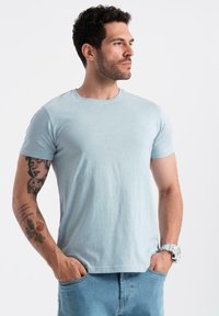 Světle modré bavlněné tričko s krátkým rukávem, kulatým výstřihem a hladkou texturou, nošené se světle modrými denimovými džíny, které ukazují viditelné tetování na paži.