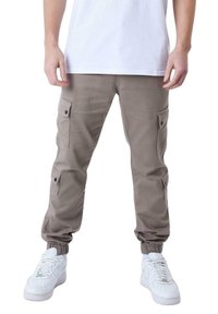 Pantalones cargo de tejido marrón claro con puños elásticos, que cuentan con múltiples bolsillos laterales y cierres a presión negros, combinados con zapatillas blancas.