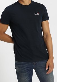 Camiseta de algodón azul marino con mangas cortas, escote redondo y un pequeño logo blanco en el pecho. Lleva puesta con vaqueros azules.