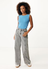 Débardeur bleu côtelé, jean gris à taille haute et jambes larges avec des coutures visibles, et baskets blanches sur un modèle devant un fond uni.