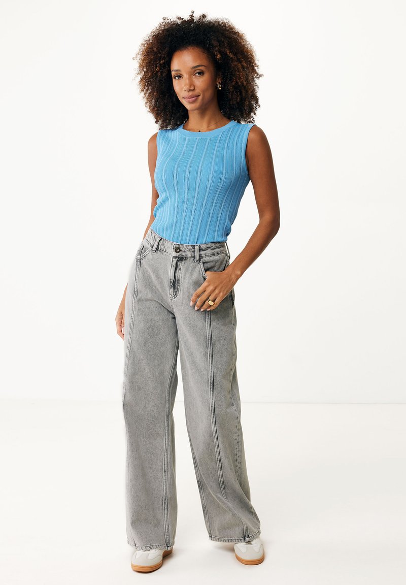 Débardeur bleu côtelé, jean gris à taille haute et jambes larges avec des coutures visibles, et baskets blanches sur un modèle devant un fond uni.