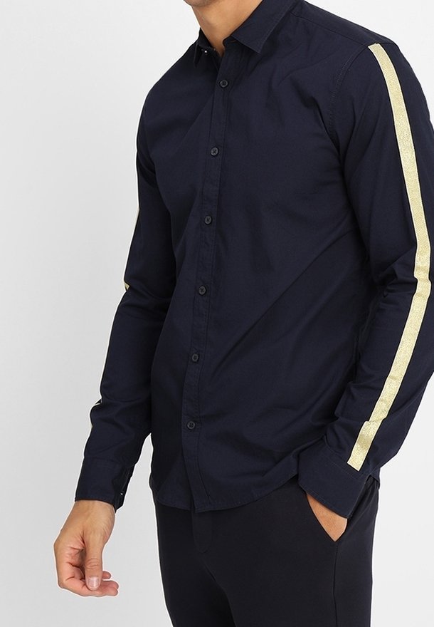 Homme portant une chemise noire cintrée à boutons avec des manches longues et une bande dorée allant de l'épaule au poignet sur les deux bras.