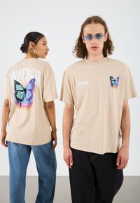 Beige suuruses T-särk, mis on valmistatud puuvillast, mille seljal on suur liblikakujutis ja vasakul ees väiksem liblikas.