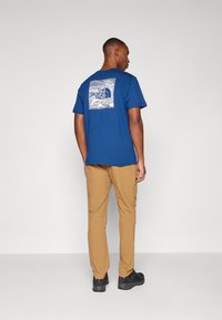 Mėlynas medvilninis T-shirts su kvadratiniu grafinu, kuriame yra "The North Face" logotipas ant nugaros, derinamas su smėlio spalvos kelnėmis ir juoda avalyne.