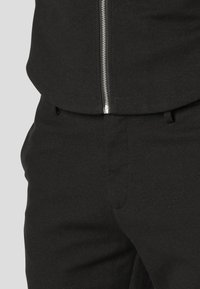 Črna zip-up jakna z gladko teksturo ima kovinski zadrgo. Usklajena je s črnimi hlačami, ki prikazujejo krojen kroj in stranske žepke.