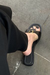 Sandalia deslizante de cuero negro con punta abierta, que presenta herrajes dorados y costuras blancas a lo largo de los bordes, colocada sobre una superficie de piedra.