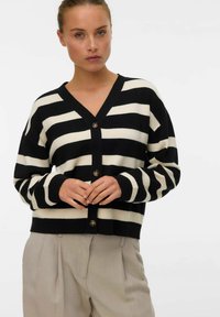Zwart en crèmekleurige gestreepte cardigan met een V-hals, knoopsluiting en lange mouwen. Draagt over lichtgekleurde broeken. Zachte, gebreide textuur.